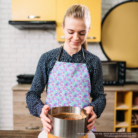 A woman wearing a Pastel Rainbow Apron by My Favourite Colour is Rainbow holds a round cake mold filled with batter in a kitchen with yellow cabinets and appliances, her colourful apron adding a splash of culinary creativity to the scene.  The apron features a soft pastel pattern of blue, pink, and white rainbow shapes.