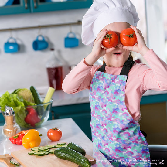 In a kitchen filled with lettuce, cucumbers, and bell peppers, a child in a chefs hat playfully holds two tomatoes over their eyes. They're wearing the Pastel Rainbow Apron by My Favourite Colour is Rainbow, which features a soft pastel pattern of blue, pink, and white rainbow shapes.