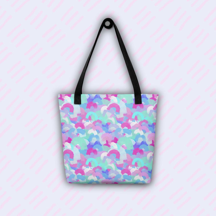 The Pastel Rainbow Tote Bag by My Favourite Colour is Rainbow features watercolour pastel rainbows in shades of blue, purple, pink, and teal.  The bag has black handles and hangs on a hook.