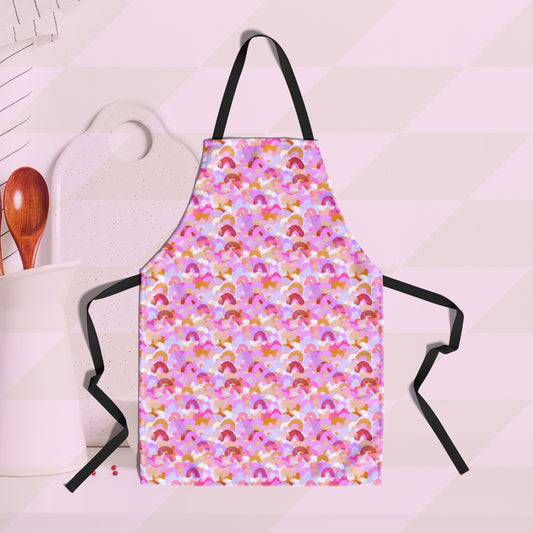 The Pink Rainbow Apron by My Favourite Colour is Rainbow features a pattern of soft watercolour rainbows in shades of pink, burgundy and gold. It hangs against a diagonal pink and white striped wall, with a white cutting board and wooden utensils ready for use.