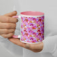 Hands hold a Pink Rainbow Mug by My Favourite Colour is Rainbow, which shows overlapping rainbow arches in pink, burgundy, and gold on a white mug with a pink interior. The person wears a white sweater and has neutral-toned nails.