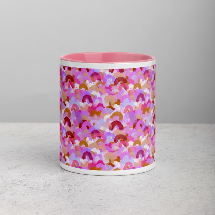 Front view of the Pink Rainbow Mug by My Favourite Colour is Rainbow featuring a pink interior and an abstract design of rainbow brushstrokes in shades of pink, burgundy, and gold. It rests on a light gray surface with a neutral background.