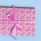 Gift box wrapped in My Favourite Colour is Rainbows Pink Rainbow Wrapping Paper Sheets with an abstract pattern  of watercolour brushstrokes resembling rainbows in shades of pink.  Shown a light blue background, tied with a bright pink ribbon forming a large bow.