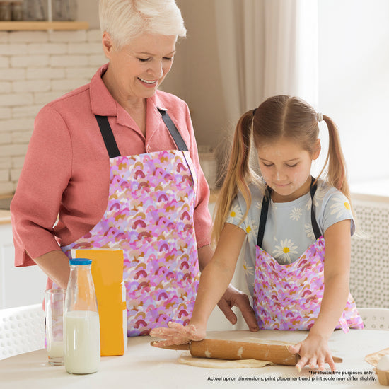 Grandmother and young girl, both wearing Pink Rainbow Aprons from My Favourite Colour is Rainbow, bake together. The girl rolls dough with a rolling pin on the table as milk and a bag of flour sit nearby. Both are focused and engaged in the activity.  The aprons feature a pattern of soft watercolour rainbows in shades of pink, burgundy and gold.