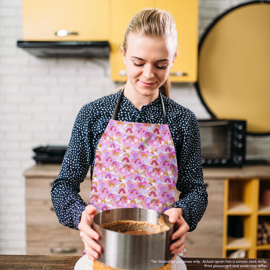 A woman wearing a My Favourite Colour is Rainbows Pink Rainbow Apron holds a pan with crumb crust in her kitchen. Smiling and admiring her creation, the kitchens yellow cabinets and white brick backsplash exude warmth and charm.  The apron features a pattern of soft watercolour rainbows in shades of pink, burgundy and gold.