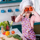 A child in a Pink Rainbow Apron from My Favourite Colour is Rainbow playfully covers their eyes with tomatoes. On the counter are cucumbers, bell peppers, and lettuce. The background features kitchenware and a plant.  The apron features a pattern of soft watercolour rainbows in shades of pink, burgundy and gold.