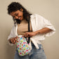 A curly-haired person smiles while adjusting the colorful Pugalicious Mini Crossbody Bag by My Favourite Colour is Rainbow. With adjustable straps, the sturdy bag complements their beige top under a white shirt and blue jeans against a plain light background.