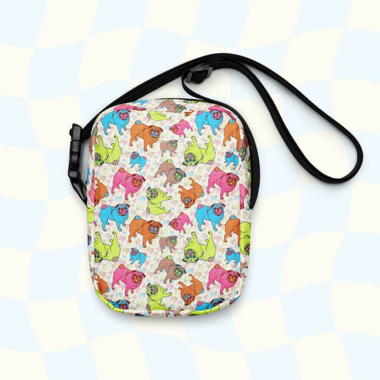 The Pugalicious Mini Crossbody Bag by My Favourite Colour is Rainbow is a small, sturdy shoulder bag with cartoon dogs on a light checkered blue-and-white background. It has adjustable black straps, a zipper closure, and offers style and water resistance.