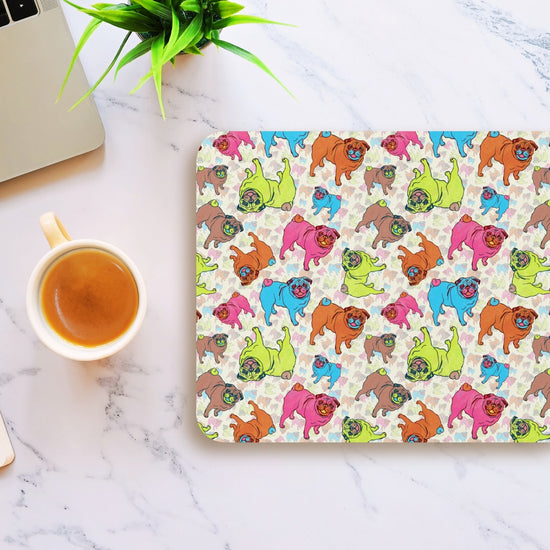 A workspace with the Pugalicious Mouse Pad by My Favourite Colour is Rainbow featuring vibrant pugs on a marble surface. Nearby are a potted plant, a laptop, and a cup of coffee.