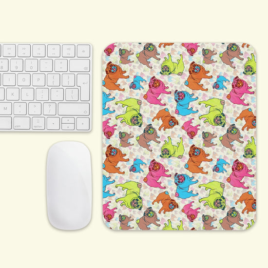 The Pugalicious Mouse Pad by My Favourite Colour is Rainbow features cartoon pugs in pink, orange, blue, green, and brown. It sits beside a white wireless keyboard and mouse on a light surface.