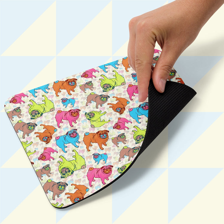 A hand lifts the Pugalicious Mouse Pad by My Favourite Colour is Rainbow, showing its vibrant cartoon pugs in pink, green, blue, and orange on a light geometrically patterned background.