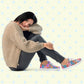 A woman sits on the floor, head resting on her knees. She wears a beige knitted sweater, black pants, and vibrant Pugalicious Canvas Sneakers from My Favourite Colour is Rainbow. The backdrop features a light design with blue stars.