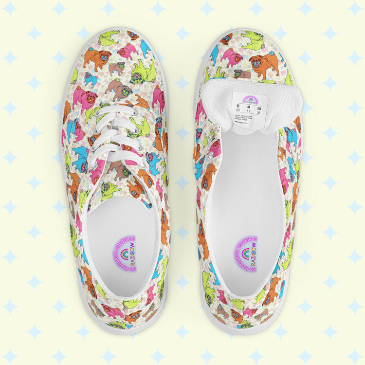 The Pugalicious Canvas Sneakers by My Favourite Colour is Rainbow showcase a delightful dinosaur pattern in green, pink, and blue on a light background. Displayed from above on a pale yellow surface with star designs, these lace-up shoes radiate charm and whimsy.