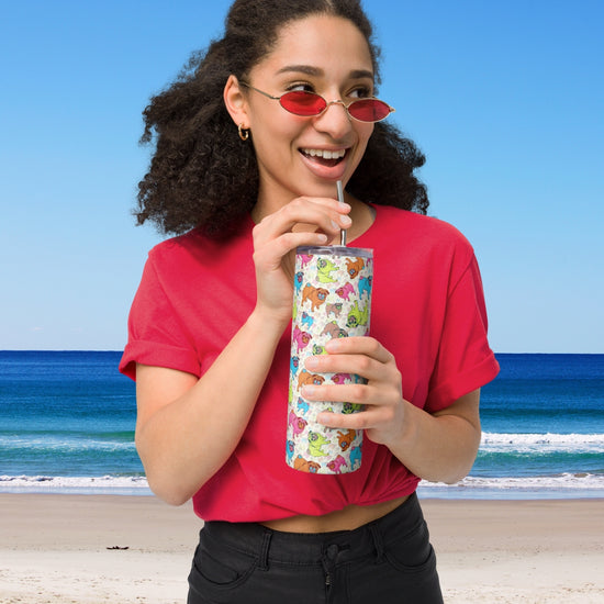 A woman in a red shirt and matching sunglasses sips from the Pugalicious Stainless Steel Tumbler by My Favourite Colour is Rainbow. Shes standing on a beach with the clear blue sky and ocean waves in the background.