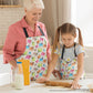 A woman and young girl, both wearing Pugalicious Aprons by My Favourite Colour is Rainbow, bake together. The girl skillfully rolls dough as the woman observes with a smile. There are a milk bottle and flour on the table.  The apron features a vibrant pattern of orange, yellow, blue and pink pugs on a light background.