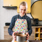 A blonde-haired person wears a colourful Pugalicious Apron by My Favourite Colour is Rainbow over a dotted shirt, concentrating on their culinary task while holding a cake ring with crust. The background features yellow cabinets and various appliances in the kitchen.  The apron features a vibrant pattern of orange, yellow, blue and pink pugs on a light background.