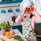 A child in a chefs hat and a Pugalicious Apron from My Favourite Colour is Rainbow stands in the kitchen, playfully holding two tomatoes over their eyes. Veggies like cucumbers and lettuce adorn the counter, with gleaming utensils in the background.  The apron features a vibrant pattern of orange, yellow, blue and pink pugs on a light background.  