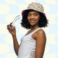 A woman smiles while wearing a Pugalicious Reversible Bucket Hat by My Favourite Colour is Rainbow, holding sunglasses. She pairs it with a sleeveless white top made from breathable fabric, standing against a light blue and white patterned background, highlighting her vibrant streetwear accessory.