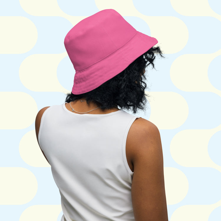 A person with curly hair wears the Pugalicious Reversible Bucket Hat by My Favourite Colour is Rainbow and a sleeveless white top. Facing away from the camera, they display their streetwear against a retro background of white and light blue ovals.