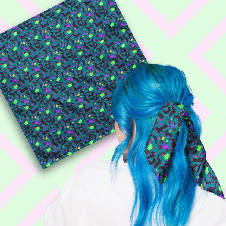 Two views of the 'Silly Sausages' Bandana by My Favourite Colour is Rainbow.  At top left, a flat lay of the bandana.  At bottom right, a person with vibrant blue hair is seen from behind, wearing the bandana in her hair.  The bandana features a fun print of black-and-magenta sausage dogs wearing bright green and purple jumpers, on a dark teal green background.