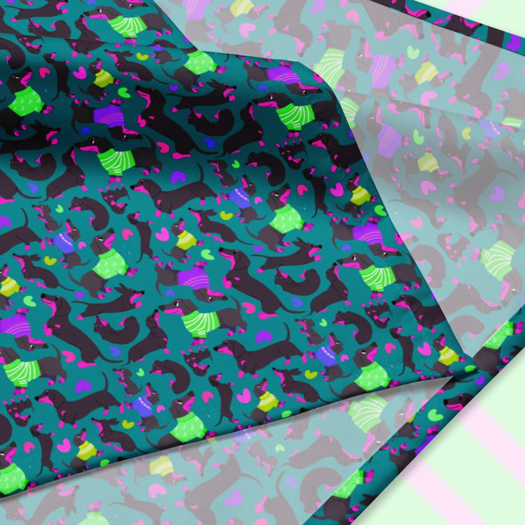 A closeup of the 'Silly Sausages' Bandana by My Favourite Colour is Rainbow featuring a fun print of black-and-magenta sausage dogs wearing bright green and purple jumpers, on a dark teal green background. The design is printed on only one side of the bandana.
