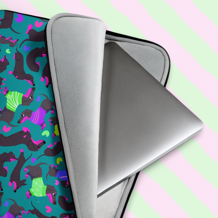 A silver laptop peeks from the Silly Sausages Laptop Sleeve – Green by My Favourite Colour is Rainbow, featuring black dachshunds in pink and green sweaters on a vibrant wavy pink and green background. It has a padded grey interior.  The print is on only one side of the laptop sleeve, the other side is black.