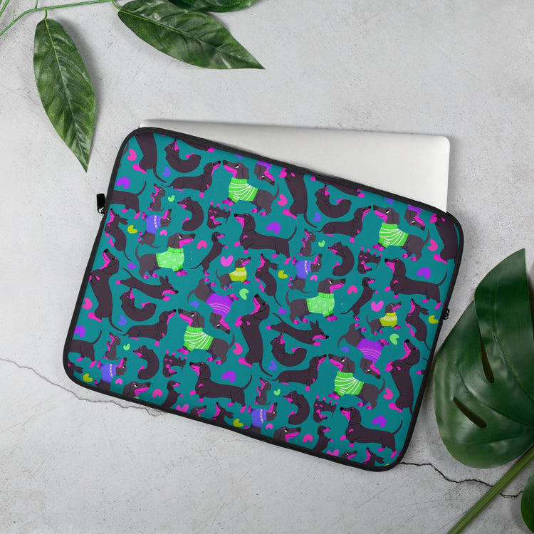 The 15 inch Silly Sausages Laptop Sleeve – Green by My Favourite Colour is Rainbow, features playful dachshunds in green and purple on a dark green backdrop with matching coloured hearts. A silver laptop is coming out of the sleeve.  Shown on a concrete background, surrounded by lush leaves.