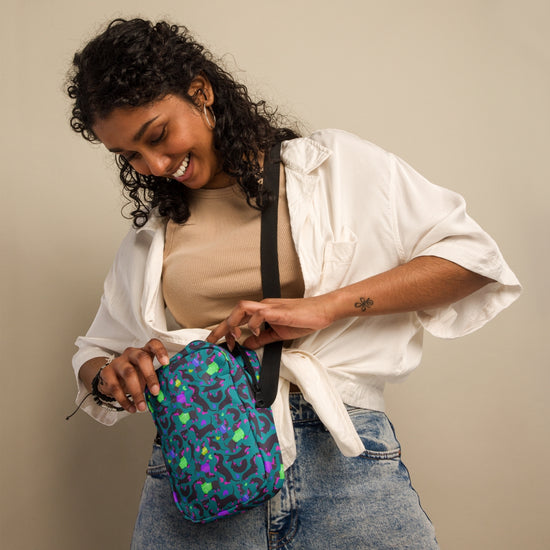 A smiling woman with curly hair in a white shirt and blue jeans holds the Silly Sausages Mini Crossbody Bag by My Favourite Colour is Rainbow. She admires the colorful bag with an adjustable strap while standing against a plain beige background.