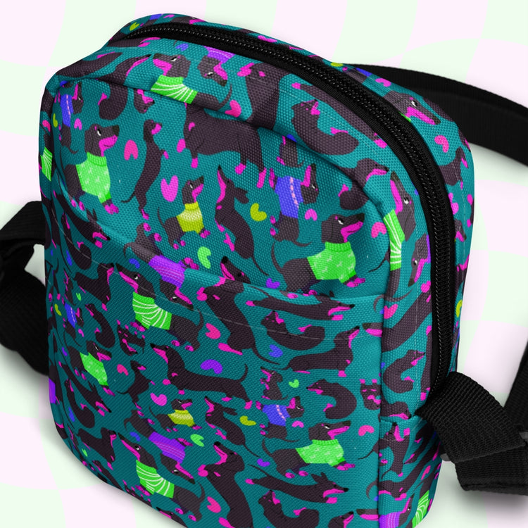 The Silly Sausages Mini Crossbody Bag by My Favourite Colour is Rainbow features a vibrant pattern with teal, purple, yellow, and black abstract shapes on a subtle light green and pink checkerboard background. With adjustable black straps and water resistance, its perfect for stylish people on the go.