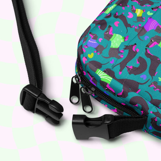 Close-up of the Silly Sausages Mini Crossbody Bag from My Favourite Colour is Rainbow, showcasing black zippers and adjustable straps. It features colorful, abstract shapes in teal, pink, and purple on a pale green and pink checkered background.