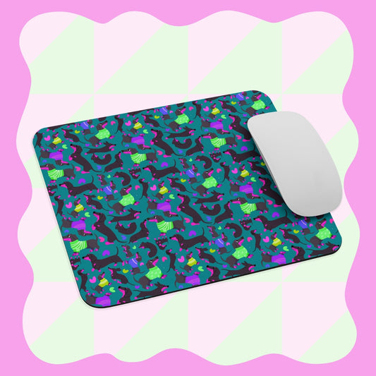 A white computer mouse sits on the Silly Sausages Mouse Pad by My Favourite Colour is Rainbow, showcasing cartoon dinosaurs in green, blue, pink, and purple on a light green and pink checkered background with a wavy lavender border.