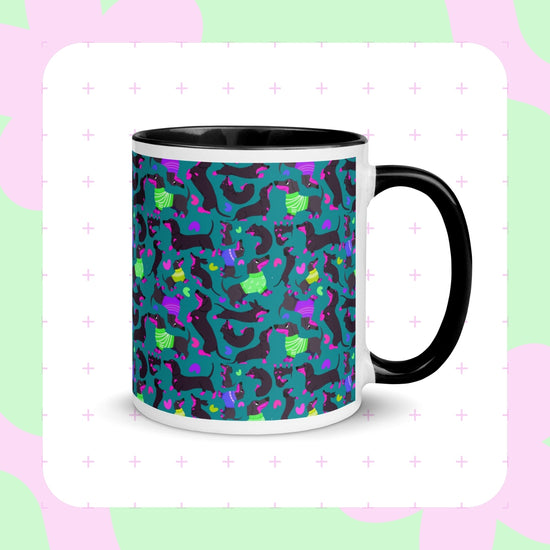 The Silly Sausages Mug by My Favourite Colour is Rainbow is a white mug with a black handle, featuring playful black-and-pink dachshunds wearing in green and purple jumpers on a dark teal background.  The mug is shown on a pink and green abstract background.