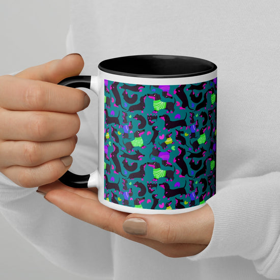 Hands holding a Silly Sausages Mug by My Favourite Colour is Rainbow; it has a black interior and features playful black-and-pink dachshunds wearing in green and purple jumpers on a dark teal background