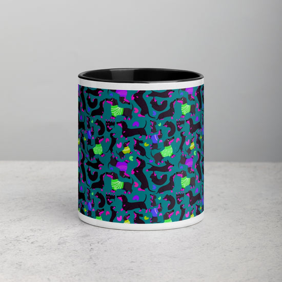 Front view of the Silly Sausages Mug by My Favourite Colour is Rainbow featuring  playful black-and-pink dachshunds wearing in green and purple jumpers on a dark teal background.  The mug has a black  interior and sites on a light grey bench.