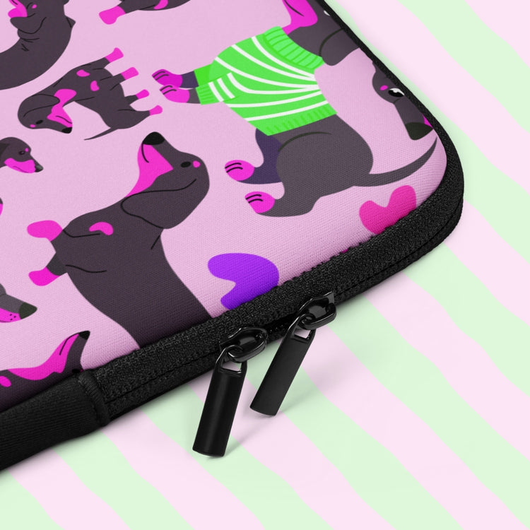 A close-up of the Silly Sausages Laptop Sleeve by My Favourite Colour is Rainbow featuring cartoon dachshunds in various poses, some donning green striped sweaters on a pink background. It includes a double zipper and a padded zipper binding.  The print is on only one side of the laptop sleeve, the other side is black.