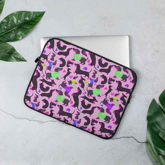 A laptop peeks out from the 13 inch Silly Sausages Laptop Sleeve – Pink by My Favourite Colour is Rainbow, showcasing a fun dachshund pattern. This scratch-proof, splash-resistant sleeve sits on a gray surface surrounded by green leaves.