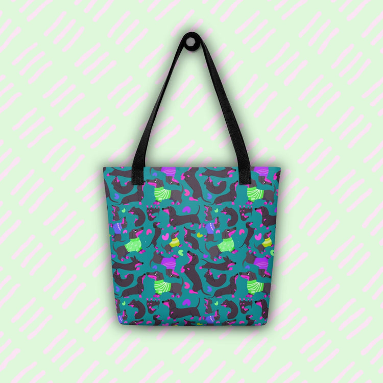 The image features the Silly Sausages Tote Bag by My Favourite Colour is Rainbow, with black straps and a fun pattern of black-and-pink dachshunds wearing green and purple jumpers on a dark teal background.