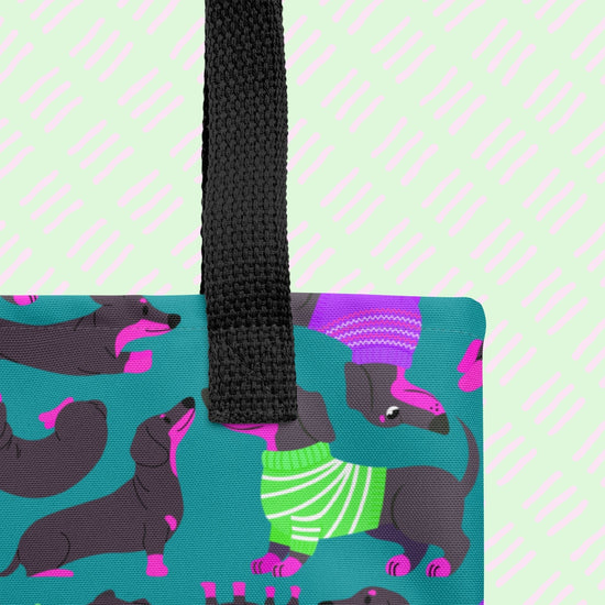 A close-up of the Silly Sausages Tote Bag by My Favourite Colour is Rainbow, featuring dachshunds in green and purple sweaters on a teal background. The tote has a black handle.