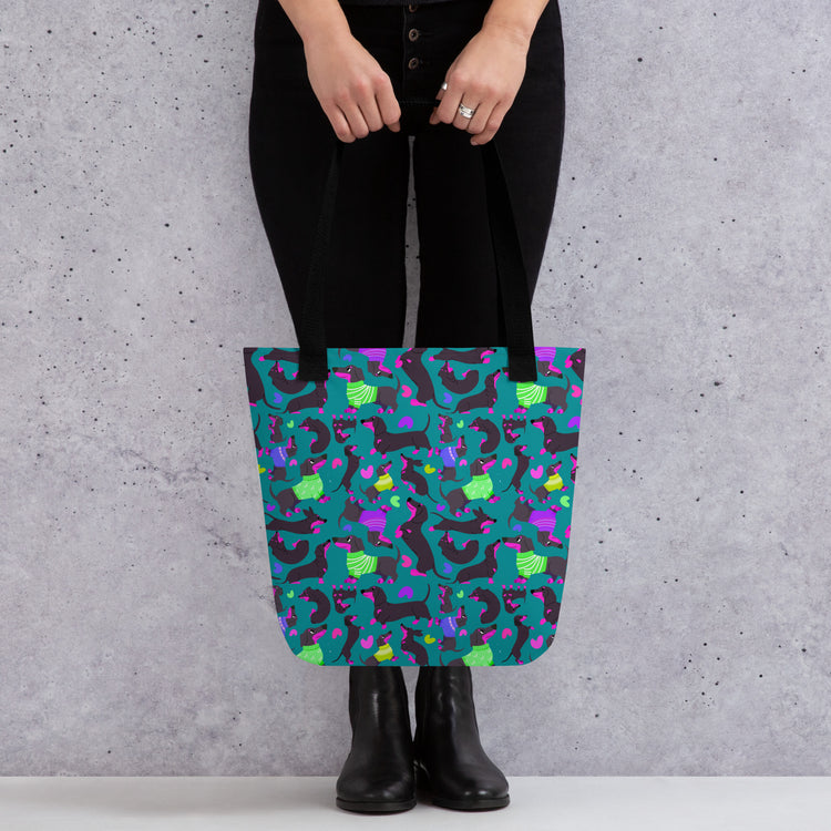 A person carries a vibrant My Favourite Colour is Rainbow Silly Sausages Tote Bag, with a fun pattern of black-and-pink dachshunds wearing green and purple jumpers on a dark teal background. They're wearing black pants and boots, standing against a textured gray wall. 