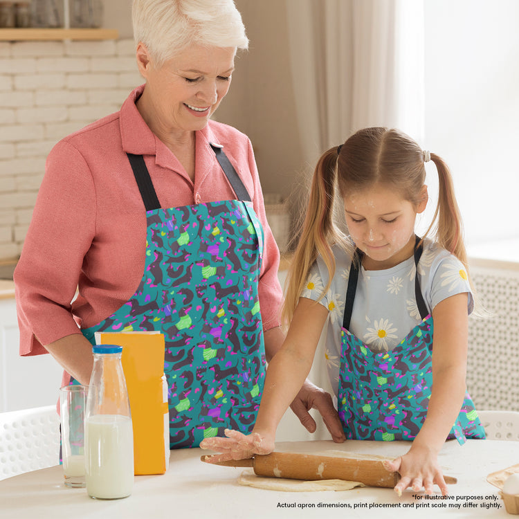 A woman and a young girl, both in Silly Sausages aprons by My Favourite Colour is Rainbow, smile while baking. The girl creatively rolls dough with a rolling pin on the table, surrounded by milk and baking supplies. The apron features a fun print of black and fuschia sausage dogs wearing purple and green jumpers on a dark green background with small hearts in matching colours. 