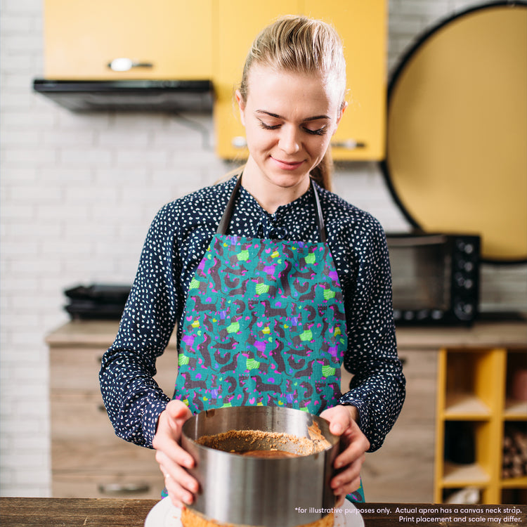 A person in a kitchen, wearing the My Favourite Colour is Rainbow Silly Sausages Apron, holds a cake pan while preparing dessert. Their culinary creativity shines as they focus on their hands and the pan, amidst an array of kitchen utensils and cabinets.  The apron features a fun print of black and fuschia sausage dogs wearing purple and green jumpers on a dark green background with small hearts in matching colours. 