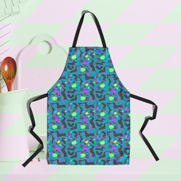 The Silly Sausages Apron by My Favourite Colour is Rainbow features a fun print of black and fuschia sausage dogs wearing purple and green jumpers on a dark green background with small hearts in matching colours.  A cutting board and utensils are in the background.