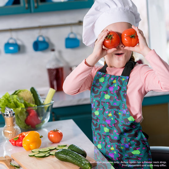 A child in a chefs hat and a Silly Sausages Apron by My Favourite Colour is Rainbow playfully holds two tomatoes in front of their eyes, standing beside a counter with cucumbers, lettuce, bell peppers, and spices, highlighting their budding culinary creativity.  The apron features a fun print of black and fuschia sausage dogs wearing purple and green jumpers on a dark green background with small hearts in matching colours. 