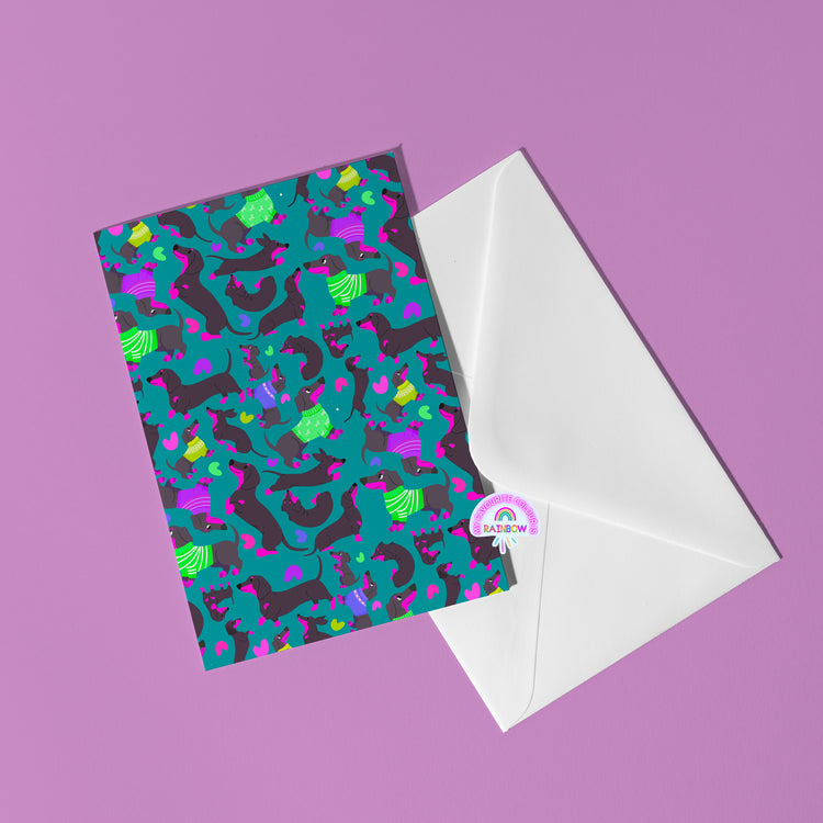 The Silly Sausages Greeting Card by My Store features playful dinosaurs and dachshunds in green, purple, blue, and pink on a teal background. It is placed on a white envelope atop a purple surface with a partial rainbow sticker visible.