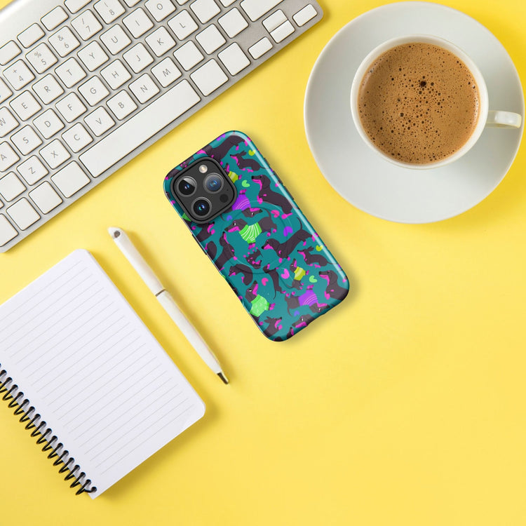 On a yellow surface, the Silly Sausages MagSafe iPhone Case by My Favourite Colour is Rainbow sits with a white keyboard, notepad with pen, and a cup of coffee on a saucer.