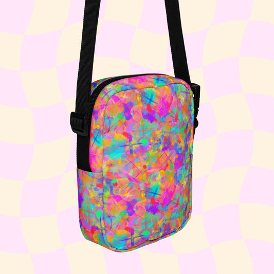 The Splotches and Crosses Mini Crossbody Bag by My Favourite Colour is Rainbow is a vibrant mini crossbody bag with a neon pink, orange, blue, and green abstract pattern. It includes an adjustable strap and black zipper on a pastel checked background.