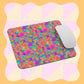 A white computer mouse sits on the Splotches and Crosses Mouse Pad by My Favourite Colour is Rainbow, featuring a vibrant abstract pattern with splashes of pink, orange, purple, and green over a pink and peach checkerboard background.