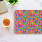 On a marble surface beside an espresso cup and partially visible laptop, the Splotches and Crosses Mouse Pad by My Favourite Colour is Rainbow displays vibrant pink, blue, green, and yellow hues. A small green plant adds a touch of nature to this abstract art setup.