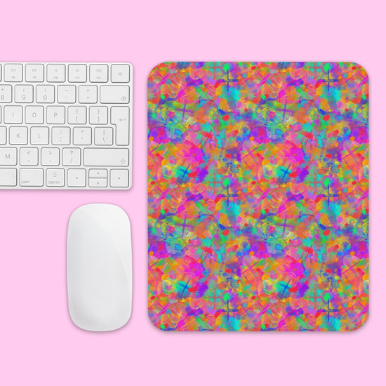 The Splotches and Crosses Mouse Pad by My Favourite Colour is Rainbow, showcasing an abstract, vibrant pattern with splashes of pink, orange, green, and blue, sits on a pink surface. To the left, theres a white wireless keyboard and mouse.