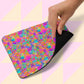 A hand lifts a corner of the Splotches and Crosses Mouse Pad by My Favourite Colour is Rainbow, featuring an abstract design in pink, blue, orange, and green on a light geometric background.